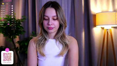 Media: Video of a young woman with fair skin and long, blonde hair, wearing a white sleeveless top, standing indoors. Background includes a lamp, green potted plant, and soft lighting.