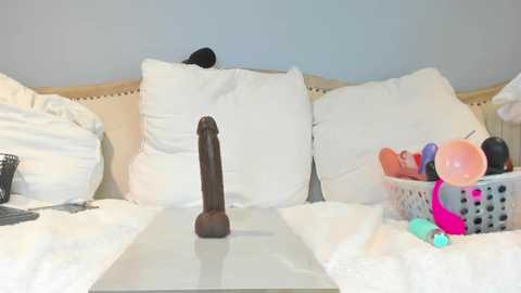 Media: Video of a neatly made bed with white pillows, a black dildo on a white sheet, and a basket of sex toys on the side.