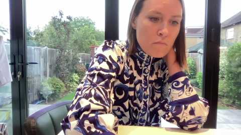 Media: Video of a woman with shoulder-length brown hair, wearing a white and blue patterned top, sitting pensively at a table by a large window with a garden view.