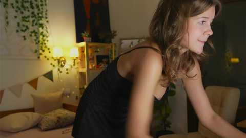 Media: Video of a young woman with wavy brown hair, wearing a black dress, leaning forward, in a cozy, dimly lit room with a fireplace, bookshelves, and greenery.