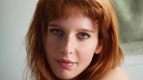 Media: Video of a fair-skinned woman with long, wavy red hair and blue eyes. She has a slight smile, wearing a white top, set against a blurred, neutral background.