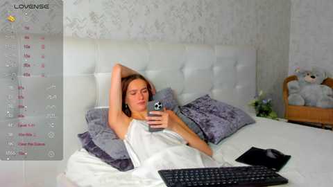 Media: Video of a young woman with light skin and brown hair, lying in a white bed with purple pillows, holding a smartphone. A plush teddy bear and a laptop are visible.