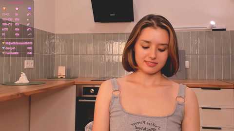 Media: A video of a young woman with straight, shoulder-length brown hair, wearing a grey tank top, standing in a modern kitchen with tiled backsplash, wooden countertops, and a flat-screen TV on the wall.