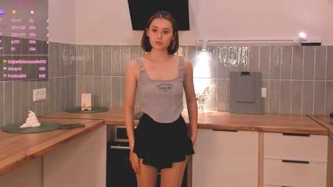 Media: Video of a young woman with fair skin and straight brown hair, wearing a light gray tank top and black skirt, standing in a modern kitchen with tiled walls and wooden countertops.