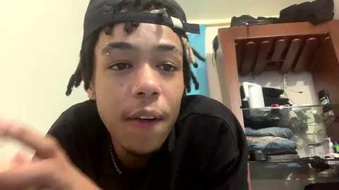 Media: Video of a young Black man with dreadlocks, wearing a black baseball cap and shirt, looking at the camera with a neutral expression. Background shows a messy closet with clothes and a mirror.