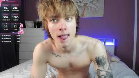 Media: Video of a shirtless, young, slender, Caucasian male with light brown hair, green eyes, and a playful expression, lying on a bed in a dimly lit room. He has tattoos on his chest and right arm.