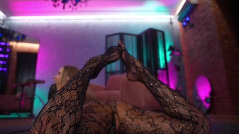 Media: Video of a blonde woman lying on her back in a dimly lit room, wearing black lace bodysuit, feet intertwined, purple and blue lights in background.