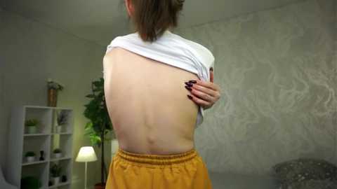 Media: Video of a fair-skinned woman lifting a white shirt to reveal her bare back, wearing a mustard-yellow skirt. She stands in a minimalistic room with a green plant, white shelves, and textured gray walls.