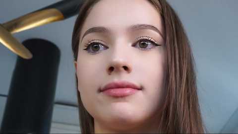 Media: Video of a young woman with fair skin, light brown hair, and subtle makeup, including winged eyeliner, glossy pink lips, and a hint of blush. She's indoors, likely in a salon or studio, with a black and gold hairdryer in the background.