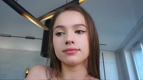 Media: Video of a young Caucasian woman with fair skin and straight brown hair, wearing minimal makeup, standing indoors with modern decor featuring a black and gold chandelier and white walls.