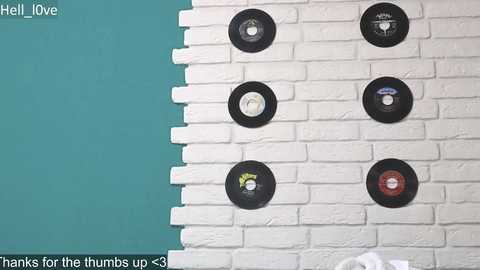 Media: Video of a white brick wall adorned with seven black record player turntables, each with unique designs. The background features a teal-painted section and a white brick section, with a digital text overlay reading \"Hells Love\" and \"Banks for the Humbus up 33.\