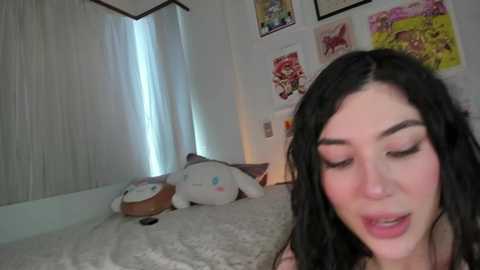 Media: Video of a young woman with long black hair, closed eyes, and a slightly open mouth, lying on a bed with a plush toy. The room has white walls with various colorful posters and a window with white curtains.