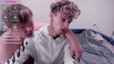 Media: Video of two young, fair-skinned, curly-haired boys with one leaning on the other, wearing white hoodies. Background shows a bed with grey blankets and a pink pillow. Screen overlay shows chat, timestamp, and emoji.