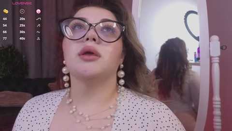 Media: Video of a plus-size woman with fair skin and brown hair, wearing glasses, pearls, and a white polka-dot top, standing in a dimly lit bedroom with a mirror reflecting her back.