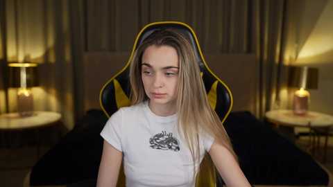 Media: Video of a young woman with long, straight blonde hair wearing a white T-shirt with a graphic design, seated in a black and yellow gaming chair in a dimly lit room with beige curtains and two lit lamps.