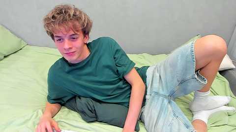 Media: Video of a young boy with curly brown hair, wearing a green t-shirt, light blue shorts, and white socks, lying on a light green bedspread.