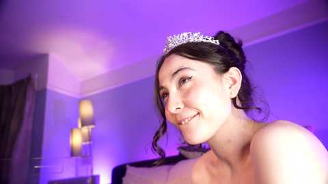 Media: Video of a smiling, fair-skinned woman with dark hair in a bun, wearing a silver tiara, in a dimly lit room with purple lighting, blurry background.