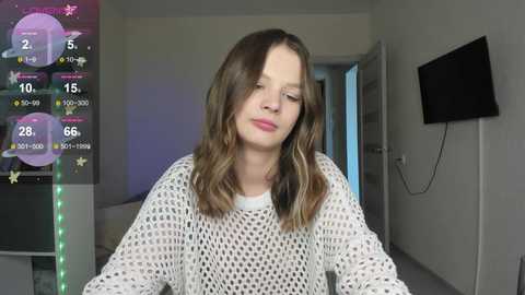 Media: A young girl with long brown hair, wearing a white crochet sweater, smiles in a modern, minimalist living room with a flat-screen TV mounted on the wall.