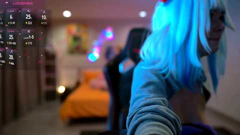 Media: Video of a vibrant bedroom with a neon blue-haired woman in the foreground, blurred background, colorful wall art, orange bed, and digital overlay displaying time and date.