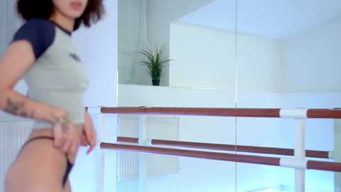 Media: Video of a woman in a gym, wearing a grey and black sports top, adjusting her black thong. She has a medium build, light skin, and curly brown hair. Background features a mirrored wall and white railings.