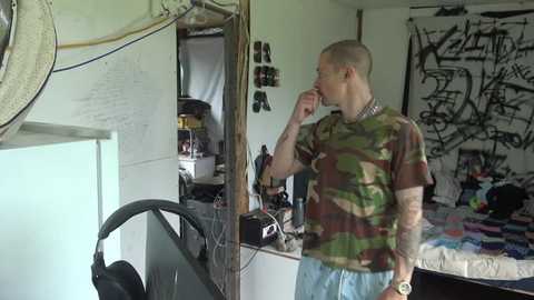 Media: Video of a bald, tattooed man in camo shirt and jeans, standing in a messy, graffiti-covered room with a bed and electronics.