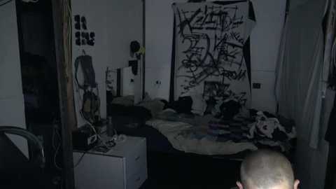 Media: Video of a dimly lit, messy bedroom with graffiti-covered wall, unmade bed, and cables on the floor. A person with short hair is visible from behind, standing near the doorway.