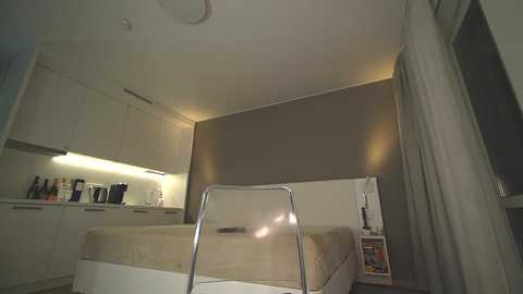 Media: A video of a minimalist, modern bedroom with beige walls and white furniture. The room features a bed against the wall, a chair in the foreground, and a small shelf with books.