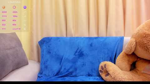 Media: Video of a cozy bedroom corner with a plush, blue blanket covering a large, brown teddy bear and a gray throw pillow on a white couch. Yellow curtains and a white wall with a sleep chart on the left.