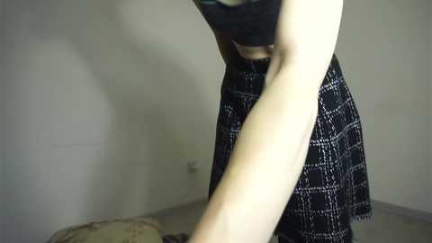 Media: A video captures a person in a black and white plaid skirt bending over, revealing a slender physique and a black bra. The background is a plain, light-colored wall and a beige carpet.