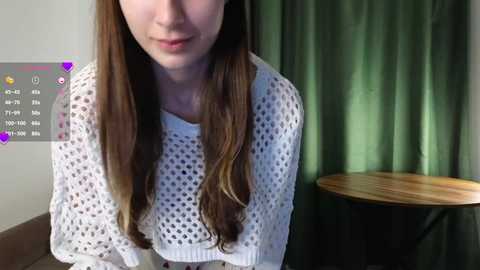 Media: Video of a young woman with long, straight brown hair, wearing a white, textured knit sweater, standing in a room with green curtains and a wooden table.