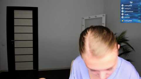 Media: Video of a man with a receding hairline, wearing a light blue shirt, standing in a minimalist room with a white wall, a potted plant, and a digital TV displaying a menu.