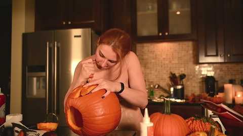 Media: Video of a redheaded, nude woman carving a large pumpkin on a kitchen counter, surrounded by lit candles, pumpkins, and kitchen appliances.