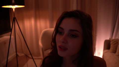 Media: Video of a young woman with dark hair and fair skin, sitting indoors with dim lighting, wearing a dark top. Background features a modern lamp and beige furniture.