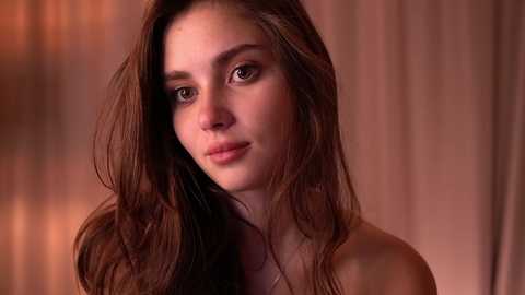 Media: A video of a young, fair-skinned woman with long, wavy brown hair and light brown eyes, wearing a strapless top, set against a blurred, warm-toned background.