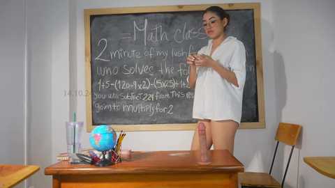 Media: Video of a young woman in a classroom, standing by a desk with a chalkboard behind her. She's wearing a white shirt, no pants, with a large, erect penis visible.