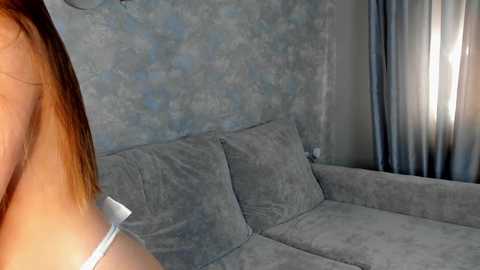 Media: Video of a beige sofa with two cushions against a textured, light blue wall, partially covered by a sheer, light blue curtain. A person's shoulder and hair are visible in the foreground.