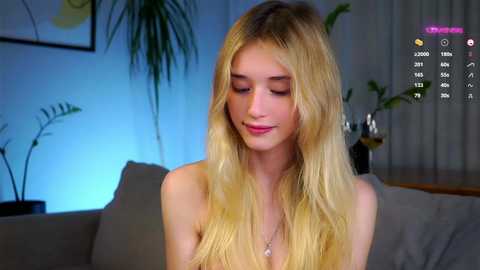 Media: Video of a topless young blonde woman with fair skin and long, wavy hair, sitting on a sofa in a modern, dimly lit room. Background includes a potted plant and a framed abstract painting.