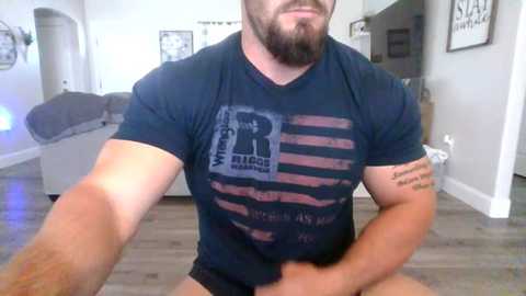 Media: Video of a muscular, bearded man with a tattoo on his arm, wearing a black t-shirt with a distressed American flag design. He's sitting on a light-colored floor in a modern, minimalist living room with white walls and wooden flooring.