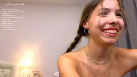 Media: A video of a smiling, topless young woman with light skin, dark hair in braids, and a silver necklace, in a cozy bedroom with a lamp and gray curtains.