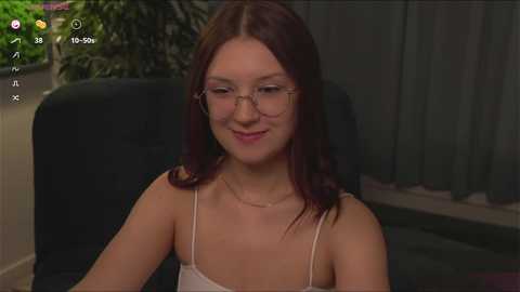 Media: Video of a young woman with long brown hair, wearing glasses, a thin spaghetti strap top, and sitting in a dark chair, indoors with greenery and curtains in the background.