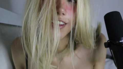 Media: Video of a fair-skinned woman with long, blonde hair covering her face, wearing a strapless top, sitting in front of a black microphone on a white background.