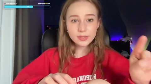 Media: Video of a young, fair-skinned woman with long brown hair, wearing a red \"WELCOME\" T-shirt, sitting in a dimly-lit room, gesturing with her right hand.