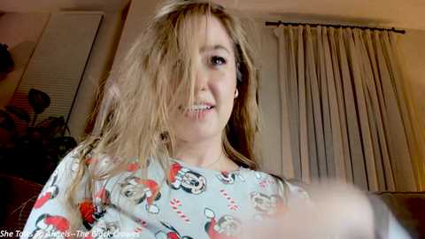 Media: Video of a young woman with long, messy blonde hair wearing a Minnie Mouse shirt, smiling slightly. Background shows beige curtains and a dimly lit room.