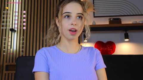 Media: Video of a young woman with curly blonde hair, wearing a purple t-shirt, seated indoors with wooden paneling, shelves, and a red heart decoration in the background.