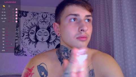 Media: Video of a shirtless young man with short brown hair, wearing a necklace, and tattoos. He gazes upward in a dimly lit room with a tapestry of sun symbols in the background.