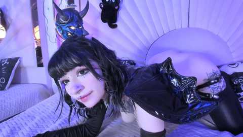 Media: Video of a pale-skinned, black-haired woman in a black latex outfit, with a horned mask, lying provocatively on a bed with white bedding. The room is dimly lit with purple accents, featuring decorative items and a plush headboard.