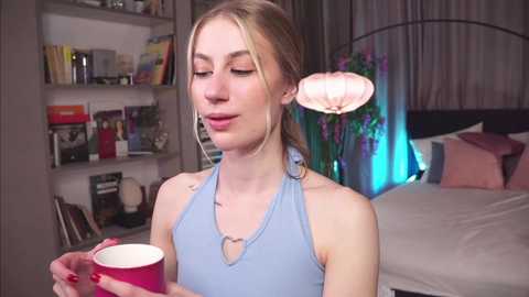 Media: Video of a young, slender woman with light skin, blonde hair, and a blue halter top, holding a pink mug, in a cozy bedroom with shelves, bed, and potted plants.