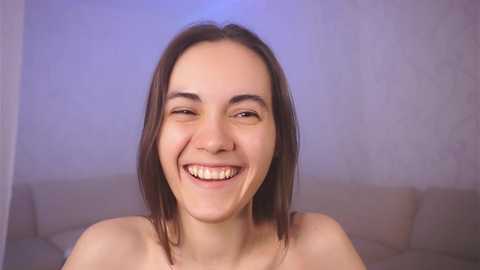 Media: Video of a smiling, light-skinned woman with straight, shoulder-length brown hair, standing topless against a soft-focus, blue-tinted background.