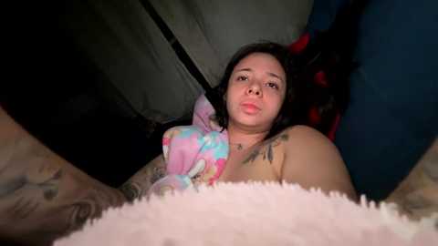 Media: Video of a topless woman with dark hair, wearing a colorful pink and blue plush toy, lying in a dimly lit tent with a blue and red patterned fabric background.