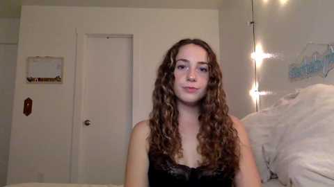 Media: Video of a young woman with curly brown hair in a black lace top, standing in a cozy bedroom with white walls, a closed door, and a bed with white sheets.